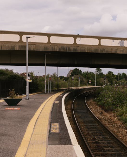 A narrow railway platform next to curved train tracks with a yellowed safety line along the edge. The platform surface features textured paving slabs and a yellow tactile strip near the edge for visua