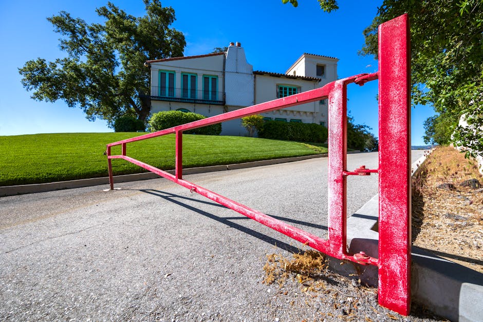 A red metal barrier gate blocks a paved pathway in front of a well-maintained front garden. The garden features a neatly mown lawn with lush, green grass and a gravel edging along the border. In the b